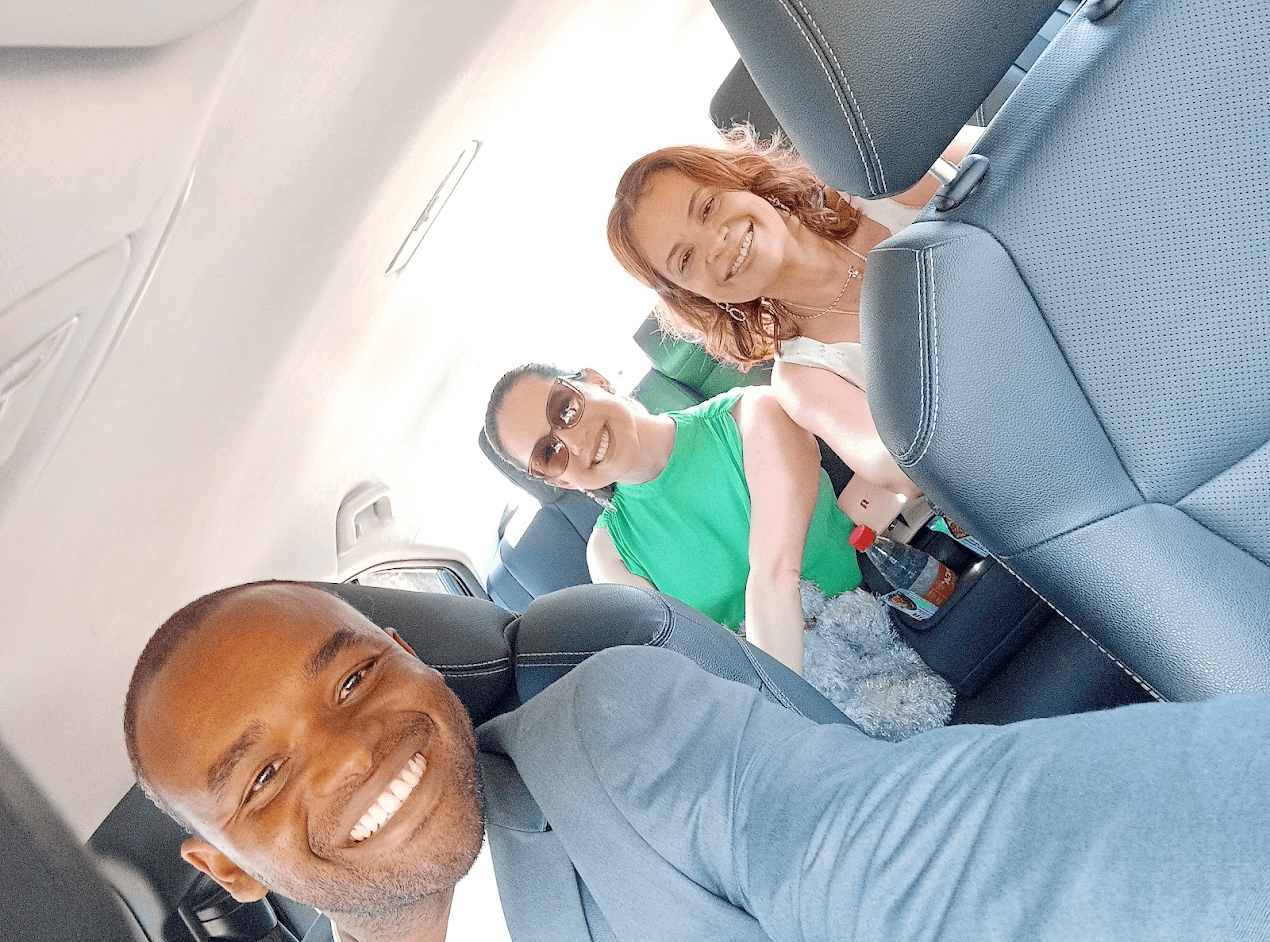 Motorista sorridente transporta duas passageiras satisfeitas em um carro executivo confortável, destacando a qualidade do serviço de transporte particular.