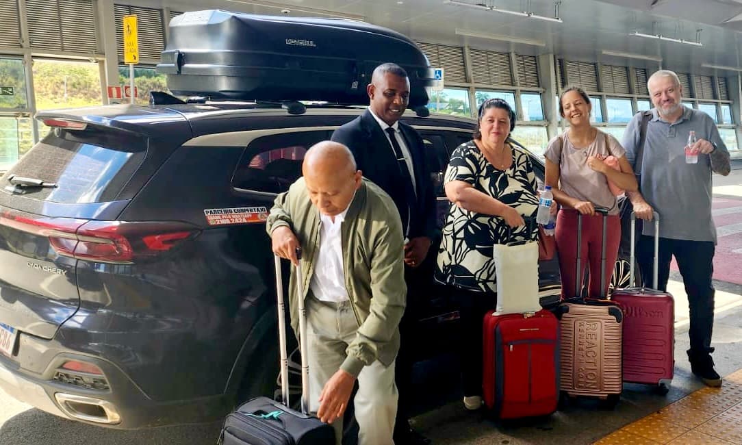 Cinco pessoas posam sorrindo ao lado de um taxi executivo, com malas de viagem na area de embarque e desembarque de um aeroporto. Incluindo um motorista uniformizado, duas mulheres e um homem com uma garrafa na mão e um homem mais velho.
